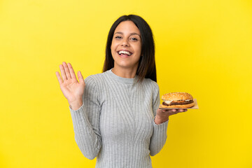 Young caucasian woman holding a burger isolated on yellow background saluting with hand with happy expression