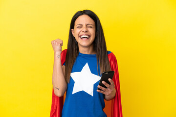 Super Hero woman isolated on yellow background using mobile phone and doing victory gesture