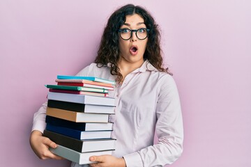 Young brunette woman with curly hair holding a pile of books afraid and shocked with surprise and amazed expression, fear and excited face.