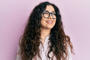 Young brunette woman with curly hair wearing casual clothes and glasses looking away to side with smile on face, natural expression. laughing confident.