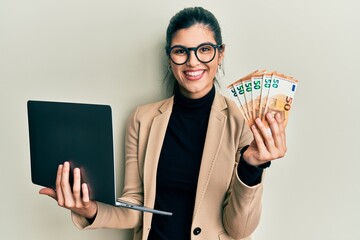 Young hispanic woman wearing business style holding laptop and 50 euros smiling with a happy and cool smile on face. showing teeth. - obrazy, fototapety, plakaty