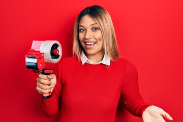 Beautiful hispanic woman holding packing tape celebrating achievement with happy smile and winner expression with raised hand