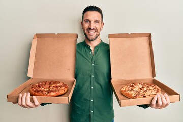 Handsome man with beard holding two italian pizzas smiling with a happy and cool smile on face....