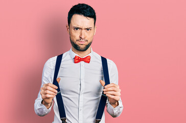 Hispanic man with beard wearing hipster look holding suspenders puffing cheeks with funny face. mouth inflated with air, catching air.