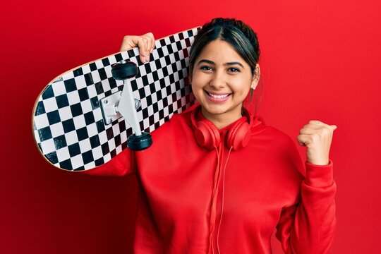 Young latin woman holding skate pointing thumb up to the side smiling happy with open mouth