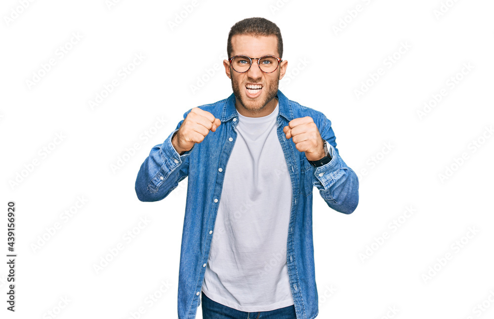 Wall mural Young caucasian man wearing casual clothes angry and mad raising fists frustrated and furious while shouting with anger. rage and aggressive concept.