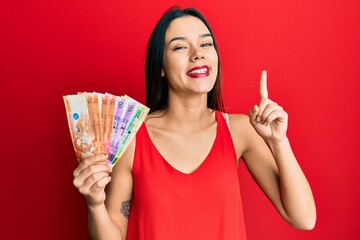 Young hispanic girl holding philippine peso banknotes smiling with an idea or question pointing finger with happy face, number one