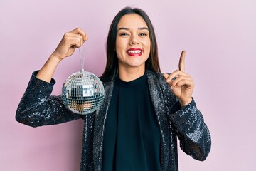 Young hispanic girl holding shiny disco ball smiling with an idea or question pointing finger with...
