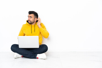 Caucasian man sitting on the floor with his laptop listening to something by putting hand on the ear