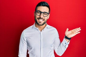 Young hispanic businessman wearing shirt and glasses smiling cheerful presenting and pointing with palm of hand looking at the camera.