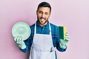 Young hispanic man wearing apron holding scourer washing dishes winking looking at the camera with...