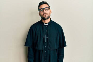 Young hispanic man wearing priest uniform standing over white background relaxed with serious expression on face. simple and natural looking at the camera.