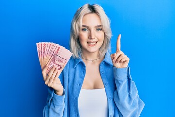 Young blonde girl holding thai baht banknotes smiling with an idea or question pointing finger with...