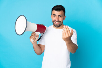 Caucasian man over isolated blue background holding a megaphone and inviting to come with hand