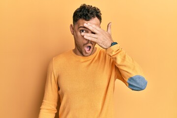 Young hispanic man wearing casual clothes peeking in shock covering face and eyes with hand,...