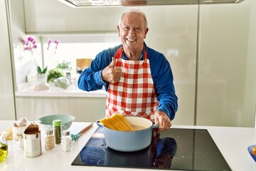 Senior man with grey hair cooking spaghetti at home kitchen smiling happy and positive, thumb up doing excellent and approval sign