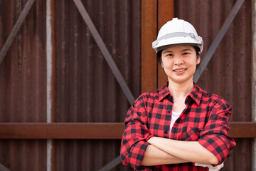 Medium shot with copy space of an Asian woman in red lumberjack shirt and safety hardhat, standing near rusty wall, smiling, crossing arms. Confident professional worker or engineer concept
