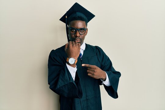Handsome black man wearing graduation cap and ceremony robe in hurry pointing to watch time, impatience, looking at the camera with relaxed expression