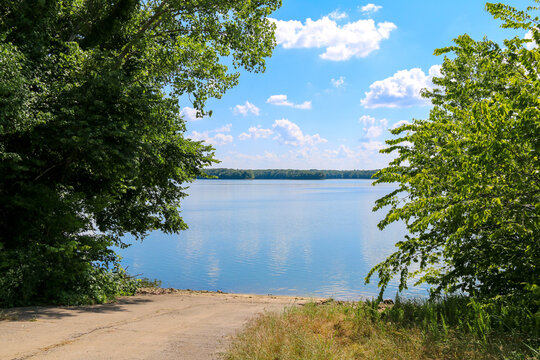 Tree Lined Lake Or River Dirt Fishing Boat Access Ramp With Reflections Under A Bright Blue Sky And Puffy White Clouds