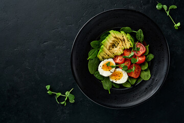 Avocado, cherry tomato, spinach and chicken egg, microgreens peas and black sesame seeds fresh salad in black bowl on black slate, stone or concrete background. Healthy food concept. Top view.