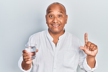 Middle age latin man drinking glass of water smiling with an idea or question pointing finger with...