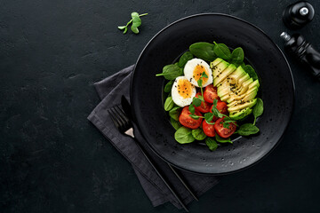 Avocado, cherry tomato, spinach and chicken egg, microgreens peas and black sesame seeds fresh salad in black bowl on black slate, stone or concrete background. Healthy food concept. Top view.