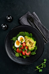 Avocado, cherry tomato, spinach and chicken egg, microgreens peas and black sesame seeds fresh salad in black bowl on black slate, stone or concrete background. Healthy food concept. Top view.