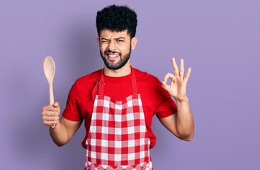 Young arab man with beard wearing baker uniform holding wooden spoon winking looking at the camera...