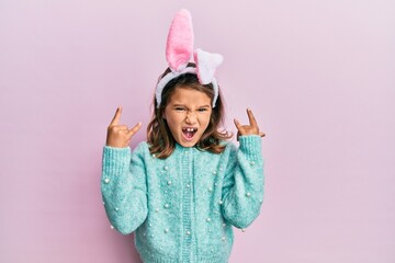 Little beautiful girl wearing cute easter bunny ears shouting with crazy expression doing rock...