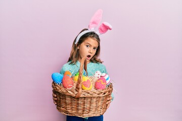 Little beautiful girl wearing cute easter bunny ears holding wicker basket with colored eggs in...