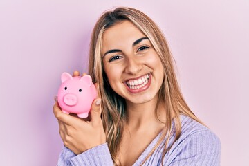 Beautiful hispanic woman holding piggy bank smiling with a happy and cool smile on face. showing teeth.