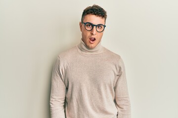 Hispanic young man wearing casual turtleneck sweater in shock face, looking skeptical and sarcastic, surprised with open mouth