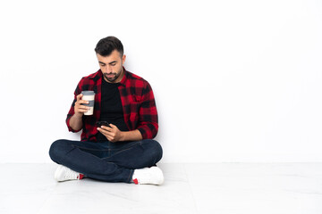 Young handsome man sitting on the floor holding coffee to take away and a mobile