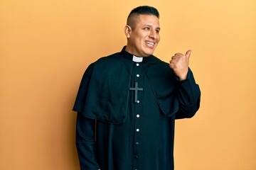 Young latin priest man standing over yellow background smiling with happy face looking and pointing...