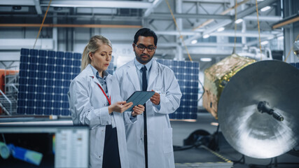 Two Engineers Talk, Use Tablet Computer while Working on Satellite Construction. Aerospace Agency...