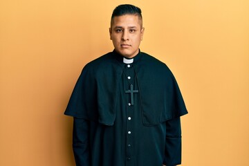 Young latin priest man standing over yellow background relaxed with serious expression on face. simple and natural looking at the camera.