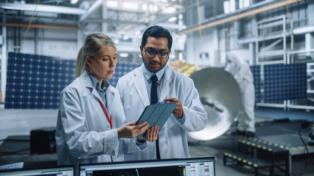 Two Engineers Talk, Use Tablet Computer While Working On Satellite Construction. Aerospace Agency Manufacturing Facility: Scientists Build, Assemble Spacecraft For Space Exploration Mission