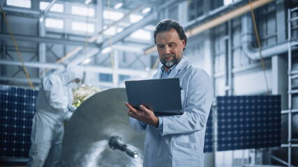 Male Engineer Uses Laptop Computer while Working on Satellite Construction. Aerospace Agency...