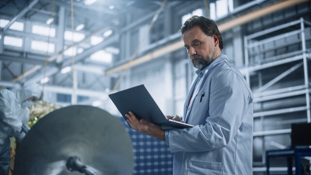 Male Engineer Uses Laptop Computer while Working on Satellite Construction. Aerospace Agency Manufacturing Facility: Scientists Develop, Assemble Spacecraft for Space Exploration, Observation Mission