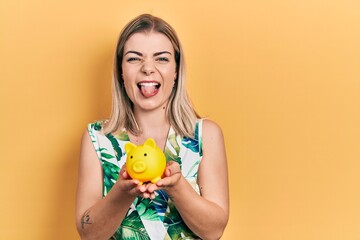 Beautiful caucasian woman holding piggy bank sticking tongue out happy with funny expression.