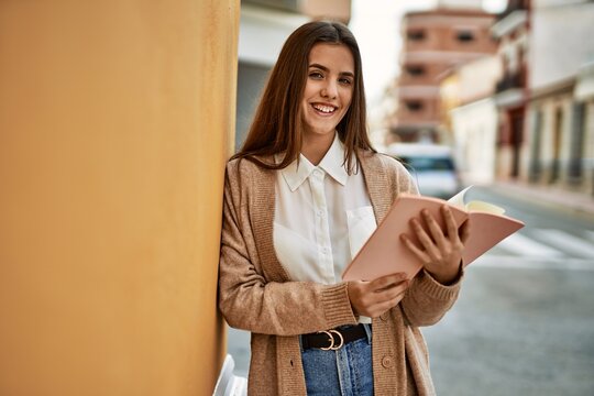 Young hispanic student girl smiling happy holding book at the city.