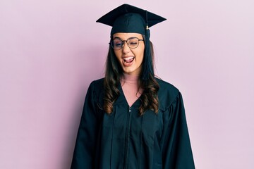 Young hispanic woman wearing graduation cap and ceremony robe winking looking at the camera with...