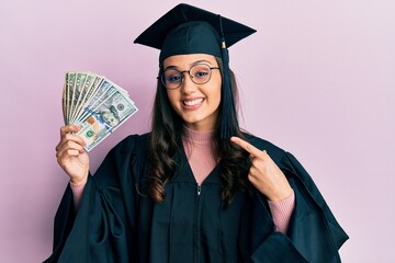 Young hispanic woman wearing graduation uniform holding usa dollars smiling happy pointing with...