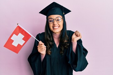 Young hispanic woman wearing graduation uniform holding switzerland flag screaming proud, celebrating victory and success very excited with raised arm