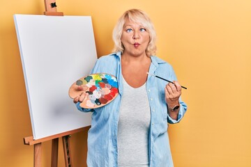 Middle age blonde woman standing drawing with palette by painter easel stand making fish face with mouth and squinting eyes, crazy and comical.