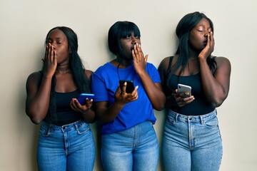 Three young african american friends using smartphone bored yawning tired covering mouth with hand. restless and sleepiness.