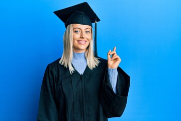 Beautiful blonde woman wearing graduation cap and ceremony robe with a big smile on face, pointing...