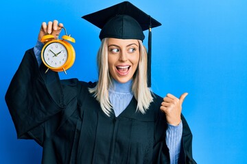 Beautiful blonde woman wearing graduation cap and ceremony robe holding alarm clock pointing thumb...