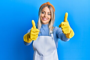 Beautiful blonde woman wearing cleaner apron and gloves approving doing positive gesture with hand,...
