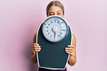Beautiful blonde woman holding weight machine to balance weight loss winking looking at the camera...